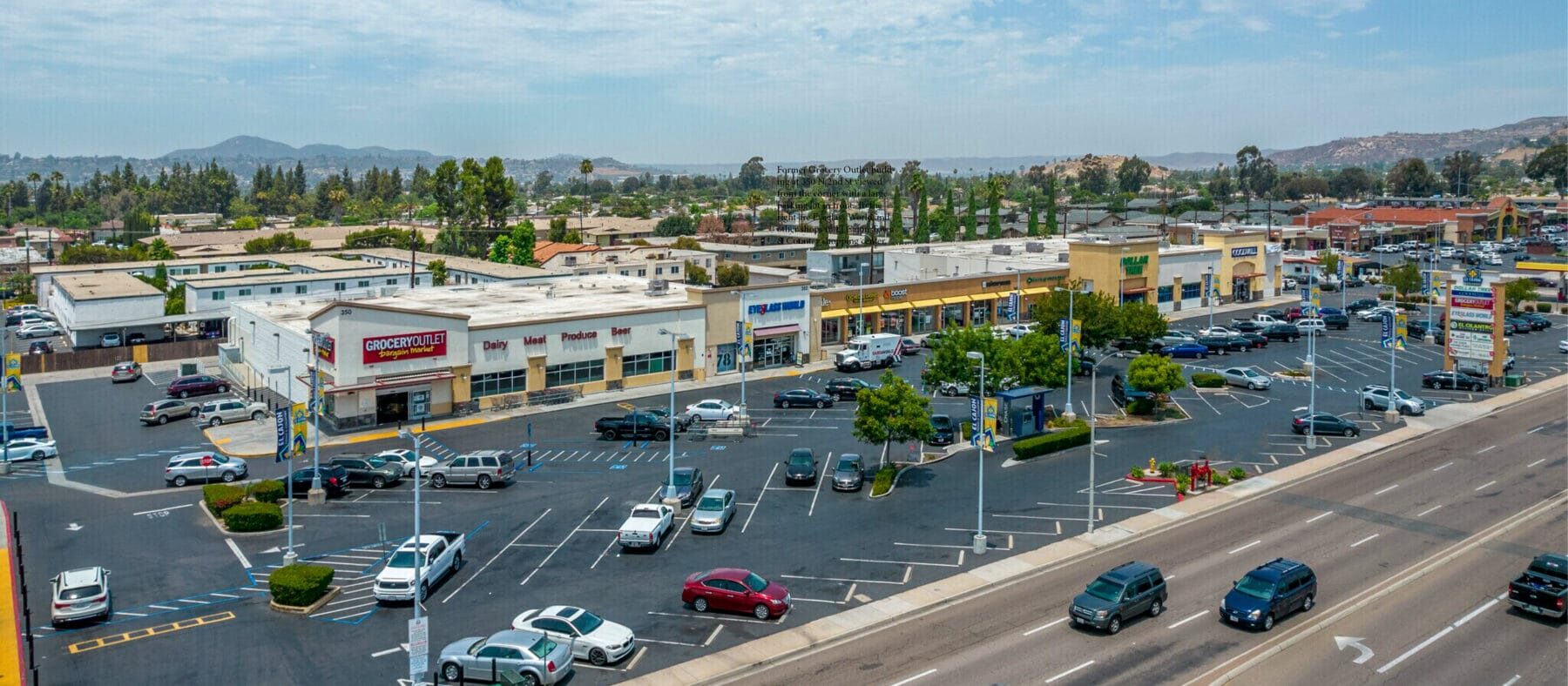 Aerial oblique of El Cajon Town & Country. The former Grocery Outlet building at 350 N 2nd St sits at the left corner, and to the right are Eyeglass World, Dollar Tree, Goodwill and other shops. A large parking lot in extends in front with N 2nd St just beyond.