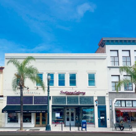 white two-story retail building, blue sky. Palm tree in front.