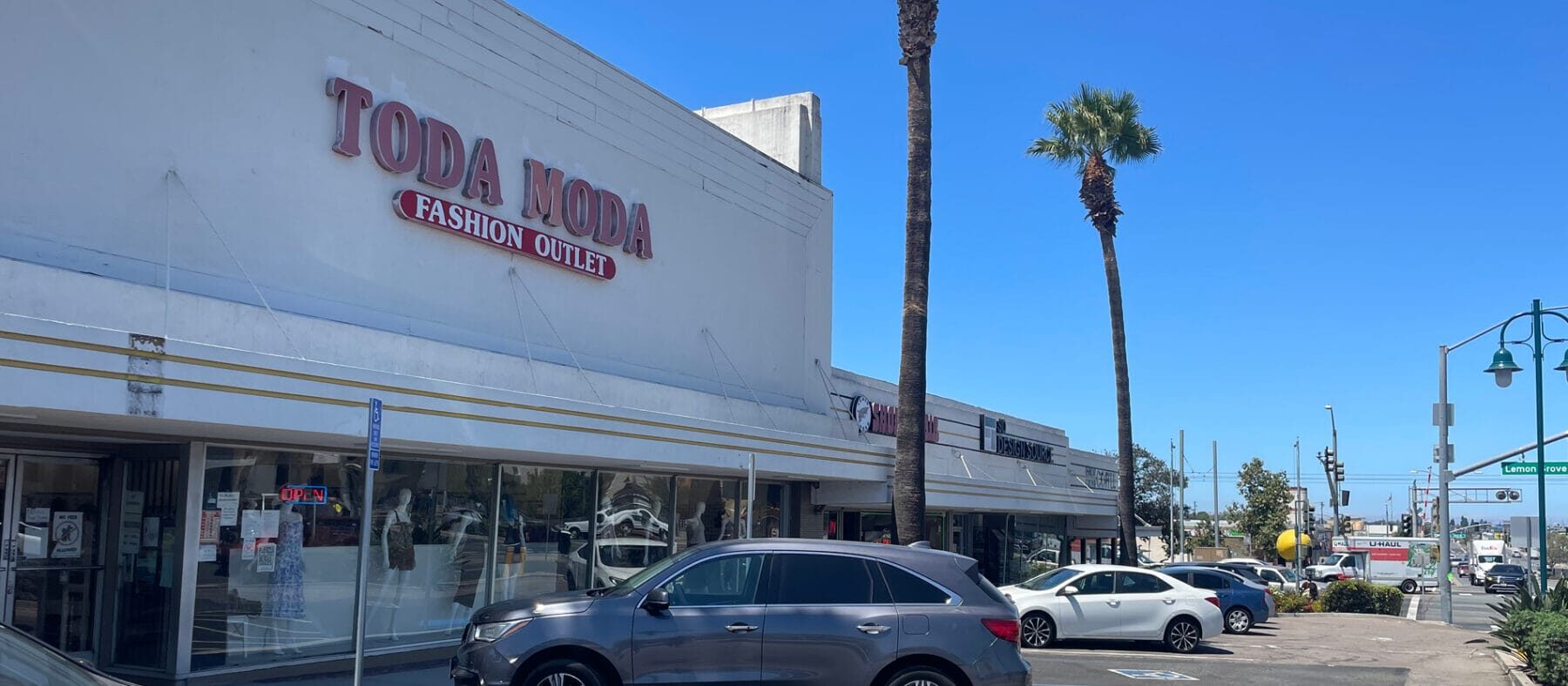 View of a single story building with multiple retailers, blue skies and palm trees in background